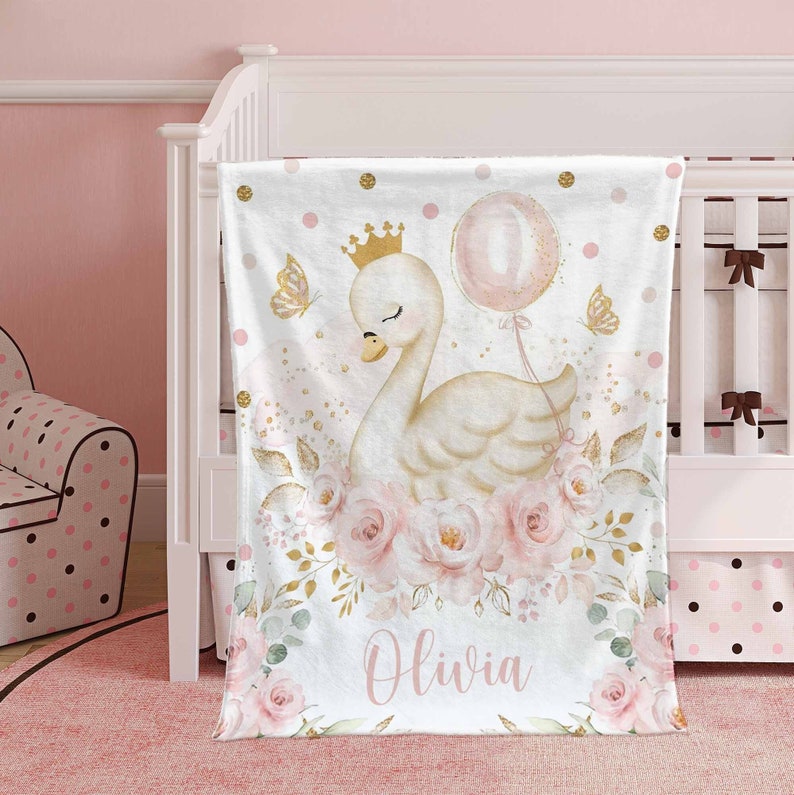 May include: A white baby blanket with a pink and gold floral design. A white swan with a gold crown and a pink balloon is in the center of the blanket. The name "Olivia" is written in pink at the bottom of the blanket.