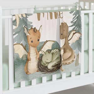 May include: A soft, white baby blanket featuring a watercolour illustration of three brown dragons. The dragons are in a woodland setting with green trees and a pale pink background. The text "Time" is written in brown letters at the top of the blanket.