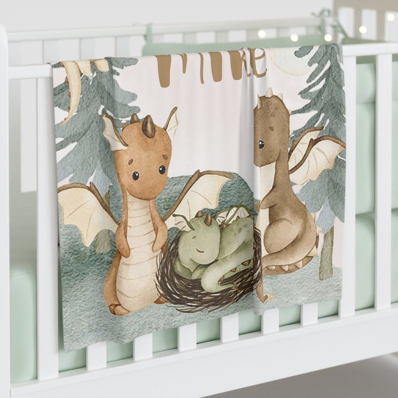 May include: A soft, white baby blanket featuring a watercolour illustration of three brown dragons. The dragons are in a woodland setting with green trees and a pale pink background. The text "Time" is written in brown letters at the top of the blanket.