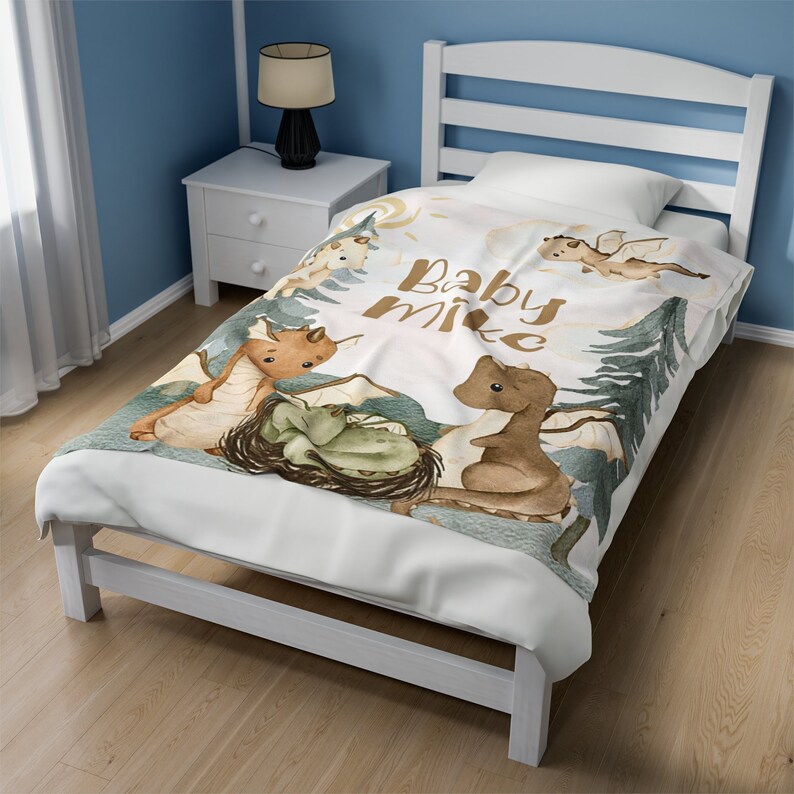 May include: A white baby blanket featuring a watercolour design of dragons and the text "Baby Mike". The blanket is on a white bed frame with a blue wall in the background.