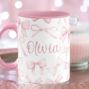 Pu&ograve; includere: Una tazza in ceramica rosa con sfondo bianco e bordo rosa. La tazza ha un motivo di fiocchi rosa e il nome "Olivia" scritto in corsivo rosa.