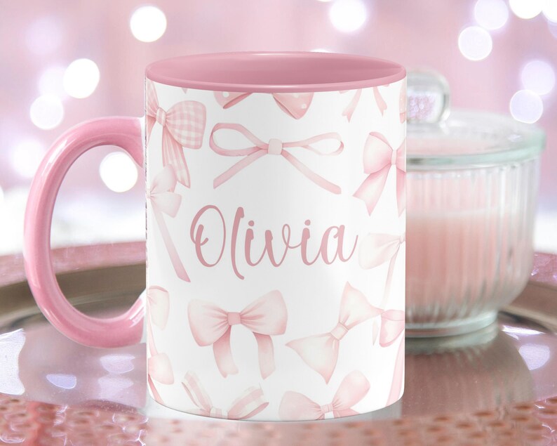 Pu&ograve; includere: Una tazza in ceramica rosa con sfondo bianco e bordo rosa. La tazza ha un motivo di fiocchi rosa e il nome "Olivia" scritto in corsivo rosa.
