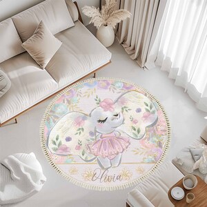 May include: A round pink and white rug with a floral design and a cartoon elephant wearing a pink tutu. The rug has a fringe border and the text "Olivia" is printed in gold lettering.