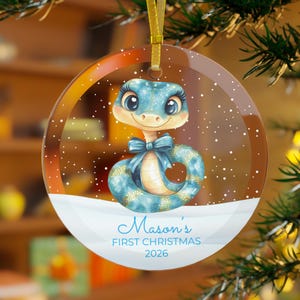 Könnte beinhalten: Ein runder, transparenter Acryl-Weihnachtsanhänger mit einem blauen Cartoon-Schlangenmotiv, großen Augen und einer blauen Schleife. Das Design zeigt weiße Schneeflocken und den Text Mason's FIRST CHRISTMAS 2026 auf einer weißen Schneefläche.