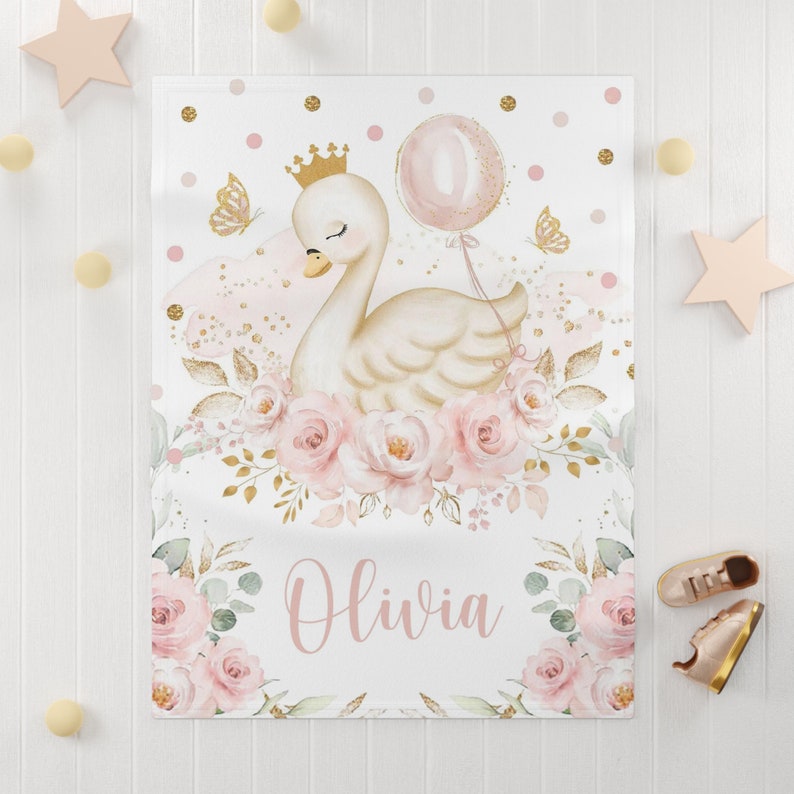 May include: A pink and gold personalized baby blanket with a white swan wearing a crown and holding a pink balloon. The blanket features a floral design with pink roses and gold accents. The name "Olivia" is written in pink script.