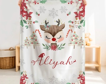 May include: A white rectangular blanket featuring a central illustration of a reindeer with a red nose, surrounded by watercolour-style Christmas trees, candy canes, snowflakes, holly, and poinsettias. The name Aliyah is printed in red script.