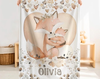 May include: A white fleece blanket featuring a watercolor illustration of an adult fox embracing a baby fox. The design includes a large heart shape, hanging heart motifs, and floral accents. The name Olivia is printed in brown lettering at the bottom.