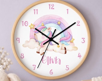 May include: A round wall clock with a light wood frame featuring a white face, black hands, and pink numbers from 1 to 12. The center displays a watercolor illustration of a unicorn with a rainbow, clouds, and stars, with the name Olivia printed below.