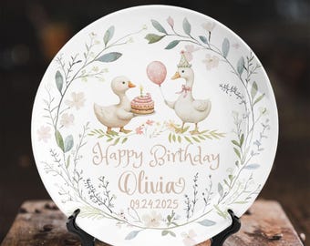 Puede incluir: Un plato decorativo blanco con una ilustración en acuarela de dos gansos, uno sosteniendo un pastel y el otro un globo. El plato está adornado con flores y follaje, y el texto "Happy Birthday Olivia 09.24.2025".