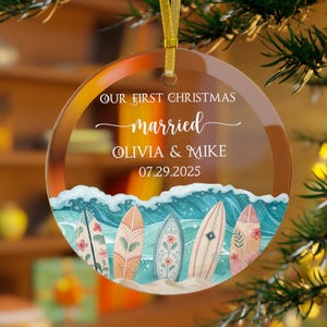May include: A clear, round Christmas ornament with a gold ribbon. The ornament features the text "OUR FIRST CHRISTMAS married OLIVIA & MIKE 07.29.2025" above a watercolor scene of surfboards and ocean waves.