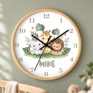Könnte beinhalten: Eine runde Wanduhr mit hellem Holzrahmen und weißem Zifferblatt, auf dem der Name MIKE in grünen Buchstaben steht. Das Design zeigt Illustrationen eines Elefantenbabys, einer Giraffe und eines Löwen auf grünem Gras mit Blättern.