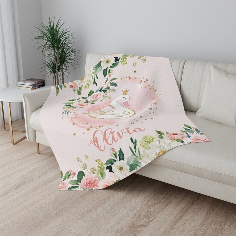 May include: Pink and white floral blanket with a gold crown and a white swan in the center. The name "Olivia" is written in gold script below the swan.