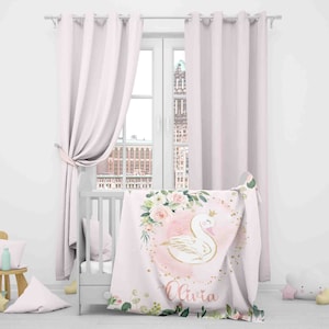 May include: A pink and white baby blanket with a gold crown, a white swan, and pink flowers. The blanket has the name "Olivia" written on it.