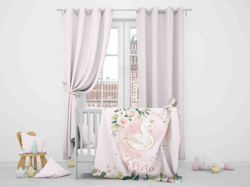 May include: A pink and white baby blanket with a gold crown, a white swan, and pink flowers. The blanket has the name "Olivia" written on it.