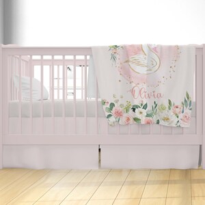 May include: A pink and white baby blanket with a gold swan design and floral accents. The blanket has the name "Olivia" written on it.