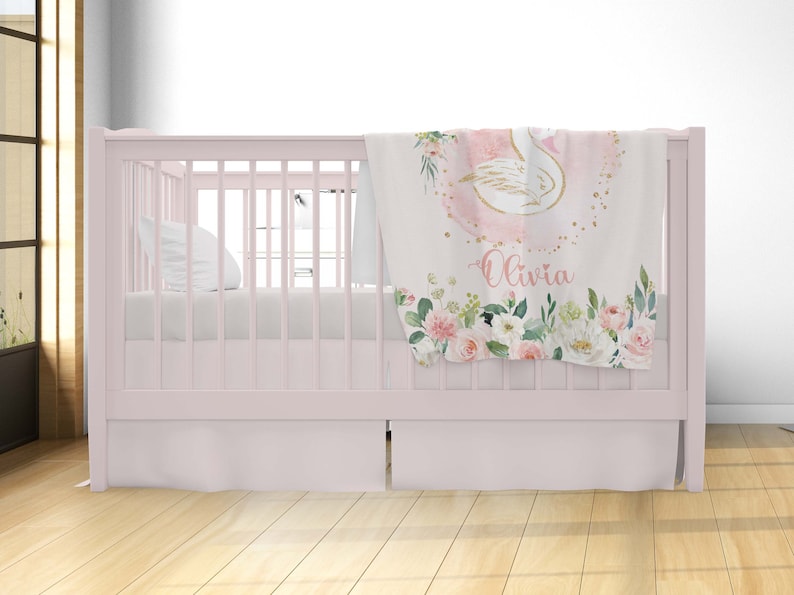 May include: A pink and white baby blanket with a gold swan design and floral accents. The blanket has the name "Olivia" written on it.