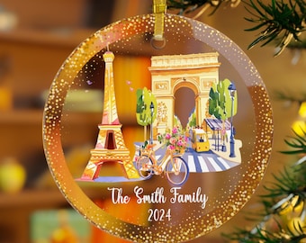 May include: A round, clear glass Christmas ornament with a gold glitter background. The ornament features a watercolor illustration of the Eiffel Tower and the Arc de Triomphe in Paris, France. The text "The Smith Family 2024" is printed on the ornament.