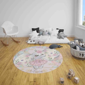 May include: A round pink and white rug with a floral design and a cartoon elephant wearing a pink tutu. The elephant's name, Olivia, is written in gold lettering on the rug.