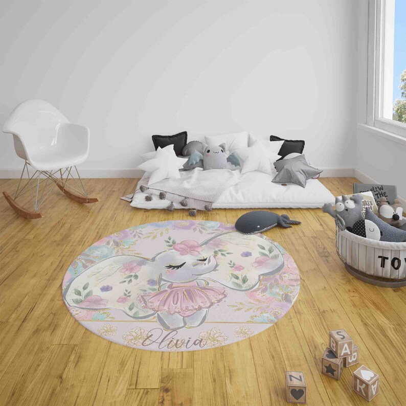 May include: A round pink and white rug with a floral design and a cartoon elephant wearing a pink tutu. The elephant's name, Olivia, is written in gold lettering on the rug.