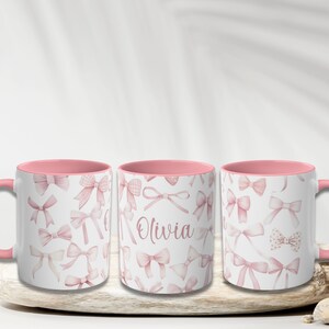 Pu&ograve; includere: Tre tazze in ceramica rosa e bianca con un bordo rosa. Le tazze sono decorate con un motivo di fiocchi rosa. La tazza centrale ha il nome "Olivia" scritto su di essa.