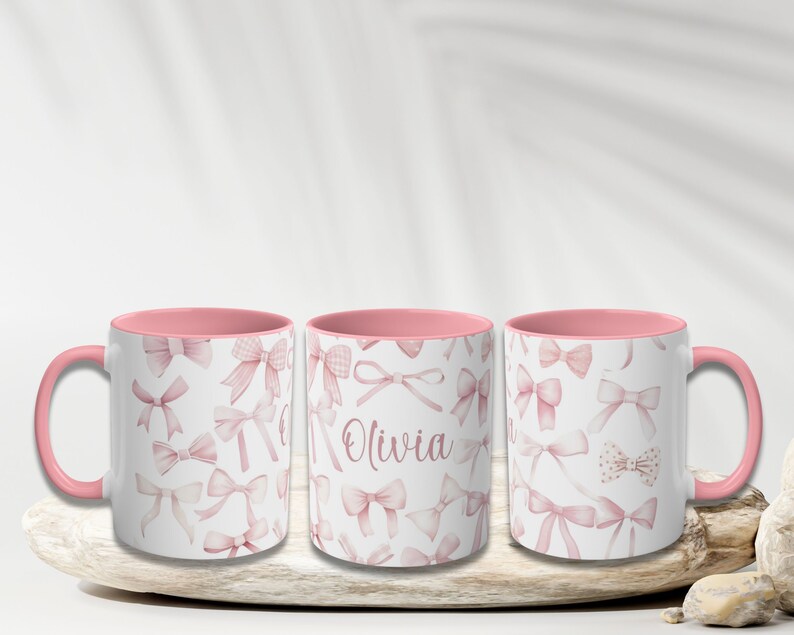 Pu&ograve; includere: Tre tazze in ceramica rosa e bianca con un bordo rosa. Le tazze sono decorate con un motivo di fiocchi rosa. La tazza centrale ha il nome "Olivia" scritto su di essa.
