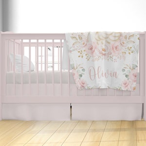 May include: A pink crib with a white crib sheet and a floral patterned baby blanket with the name "Olivia" on it.