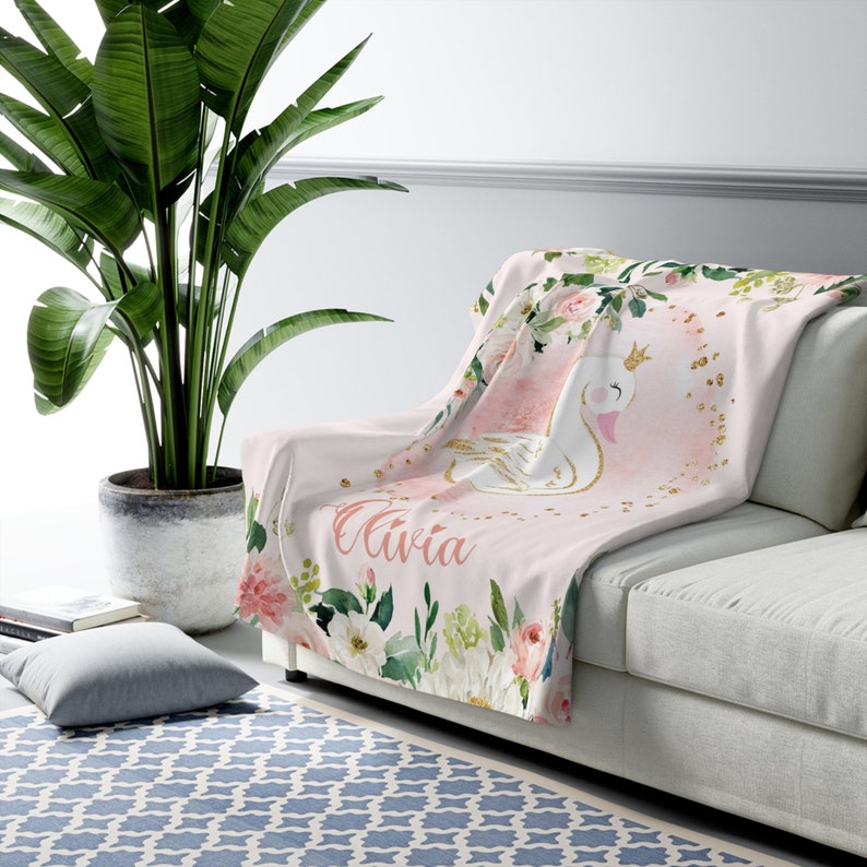 May include: Pink and green floral throw blanket with a white swan with a gold crown and gold glitter. The blanket has the name "Olivia" written on it.