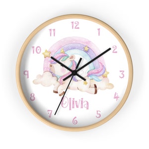 May include: A white and pink unicorn with a rainbow and stars on a white background. The clock face has pink numbers and the name "Olivia" is written below the unicorn.