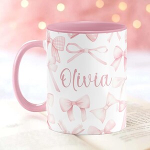 Pu&ograve; includere: Una tazza in ceramica rosa e bianca con un bordo e un manico rosa. La tazza ha uno sfondo bianco con un motivo a fiocco rosa e il nome "Olivia" stampato in rosa.