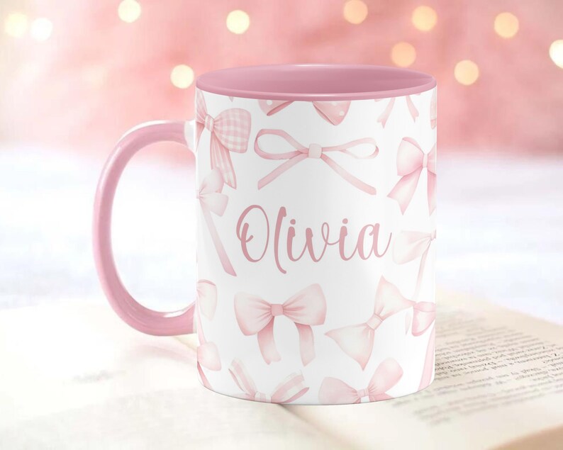 Pu&ograve; includere: Una tazza in ceramica rosa e bianca con un bordo e un manico rosa. La tazza ha uno sfondo bianco con un motivo a fiocco rosa e il nome "Olivia" stampato in rosa.