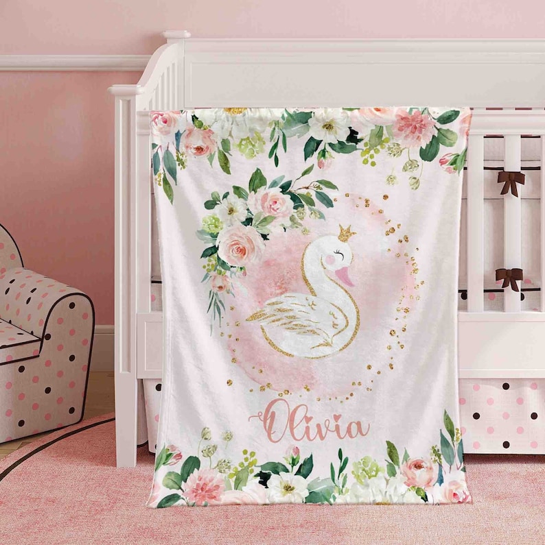 May include: A white and gold swan with a crown on its head is surrounded by pink and green watercolor flowers. The background is a light pink with gold glitter. The name "Olivia" is written in pink script at the bottom of the blanket.
