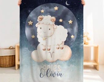 May include: A rectangular fleece blanket featuring a white cartoon ram standing on a beige cloud against a dark blue moonlit sky with yellow stars. The text Aries is printed above the name Olivia in a dark blue script font on the lower section.