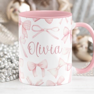 Pu&ograve; includere: Una tazza in ceramica rosa con sfondo bianco e fiocchi rosa. La tazza ha il nome "Olivia" scritto su di essa.
