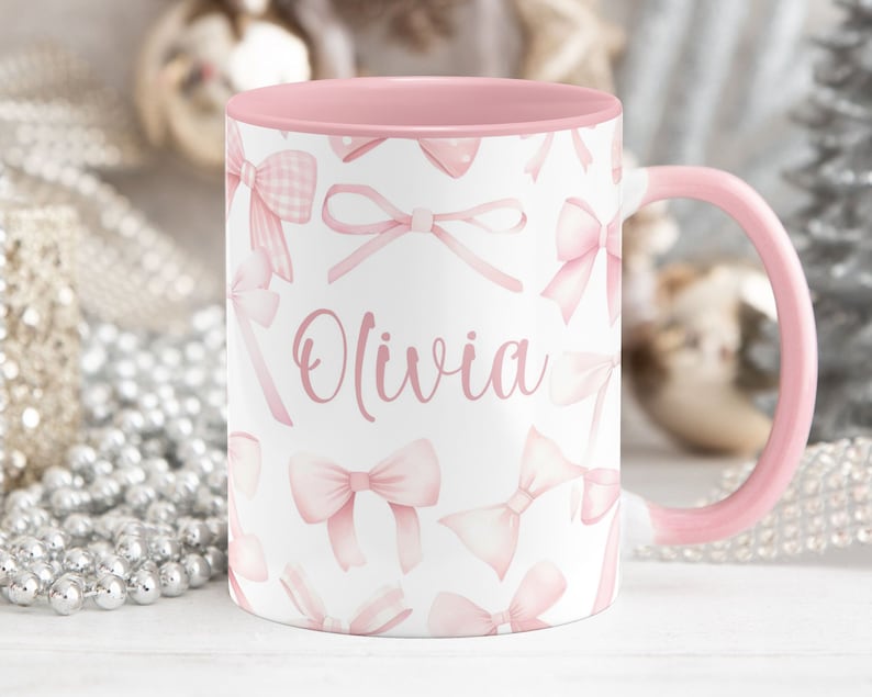 Pu&ograve; includere: Una tazza in ceramica rosa con sfondo bianco e fiocchi rosa. La tazza ha il nome "Olivia" scritto su di essa.