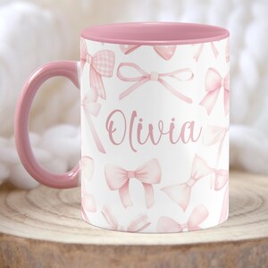 Pu&ograve; includere: Una tazza in ceramica rosa e bianca con interno rosa. La tazza ha uno sfondo bianco con un motivo a fiocco rosa e il nome "Olivia" scritto in corsivo rosa.