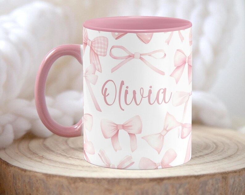Pu&ograve; includere: Una tazza in ceramica rosa e bianca con interno rosa. La tazza ha uno sfondo bianco con un motivo a fiocco rosa e il nome "Olivia" scritto in corsivo rosa.