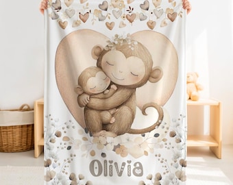 Könnte beinhalten: Eine weiße Fleecedecke mit einer großen Illustration einer Affenmutter, die ein Affenbaby vor einem beigefarbenen Herzen umarmt. Das Design umfasst florale Akzente, hängende Herzformen und den Namen Olivia in braunen Buchstaben am unteren Rand.
