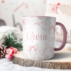 Pu&ograve; includere: Una tazza in ceramica bianca con un bordo e un manico rosa. La tazza ha un design ad acquerello rosa con fiocchi e il nome "Olivia" scritto in corsivo rosa.