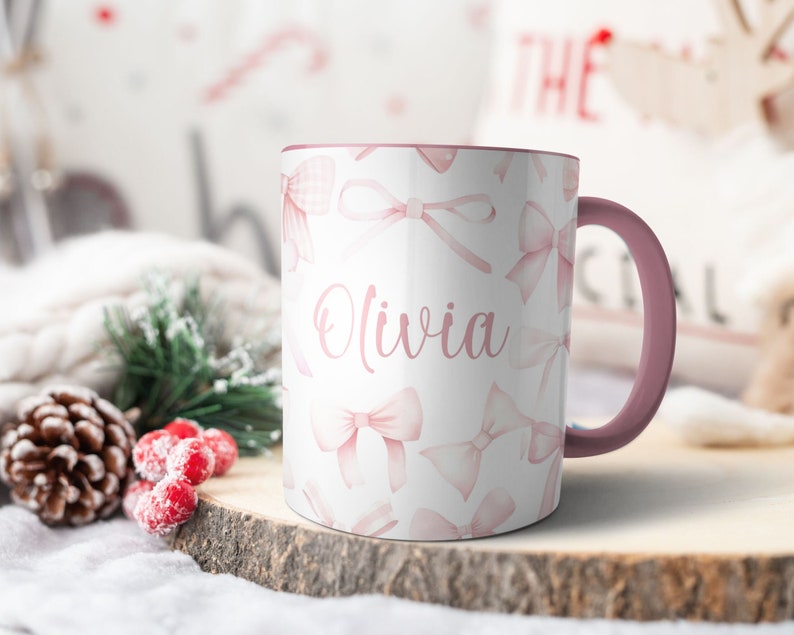 Pu&ograve; includere: Una tazza in ceramica bianca con un bordo e un manico rosa. La tazza ha un design ad acquerello rosa con fiocchi e il nome "Olivia" scritto in corsivo rosa.