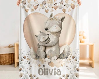 May include: A white fleece blanket featuring a large illustration of a gray wolf hugging a smaller wolf cub. The design includes a large beige heart, a border of small hearts, and floral accents. The name Olivia is printed in large gray letters.