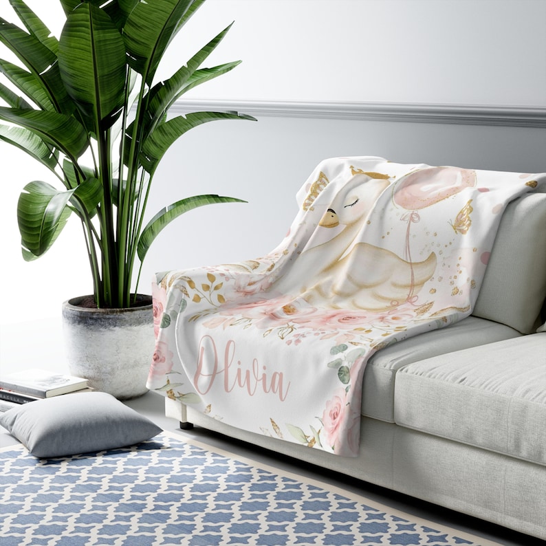May include: A white fleece blanket with a pink and gold floral design. A white swan with a gold crown and a pink balloon is featured on the blanket. The name "Olivia" is written in pink script on the blanket.