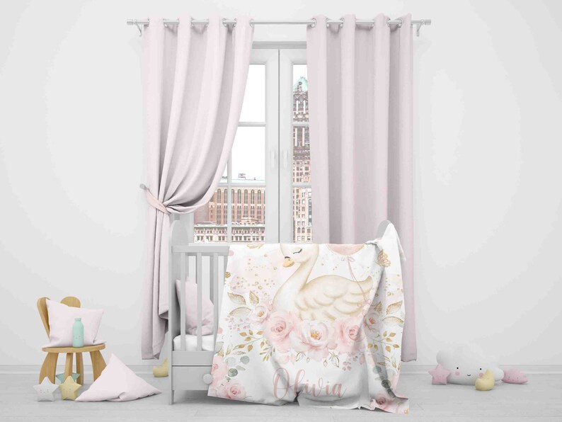 May include: A white crib with a pink and gold floral blanket featuring a swan with a balloon. The blanket has the name "Olivia" written on it.