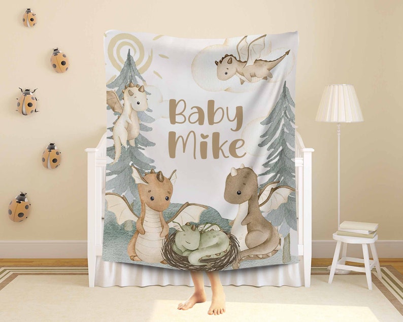 May include: A white baby blanket featuring a watercolour design of dragons and trees. The blanket has the text "Baby Mike" in brown lettering.