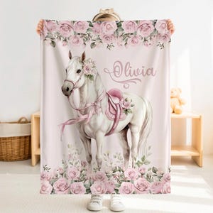 May include: A light pink blanket featuring a white horse with a pink saddle and floral accents. The blanket is decorated with pink roses and the name "Olivia" in a script font. The blanket is held up by a person.