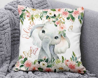 Op de afbeelding: Een wit kussen met een aquarel-illustratie van een grijze olifant die een roze bloemenkroon draagt. De olifant wordt omringd door roze en witte bloemen en groene bladeren. De naam "Olivia" staat in roze schrift op het kussen.