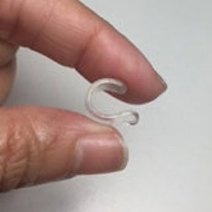 May include: A clear, plastic C-shaped clip held between fingers. The clip has a curved design with a small hook at each end. The clip appears to be small and lightweight.