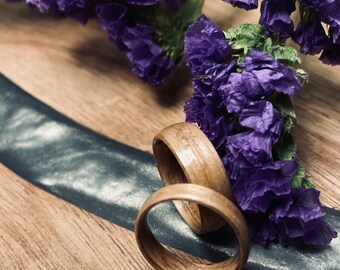 May include: Two wooden rings stacked on top of each other. The rings are a light brown color and have a natural wood grain. The rings are sitting on a wooden surface with a dark blue and silver inlay. Purple flowers are in the background.