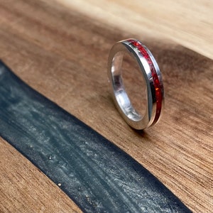 May include: A silver ring with a thin red inlay. The ring is on a wooden surface.