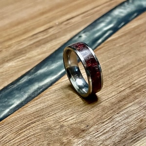 May include: A stainless steel ring with a wide band. The ring has a vibrant red and white speckled inlay. The ring is set against a wooden surface with a dark grey accent.