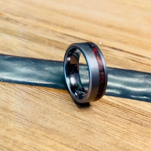 May include: A gray titanium ring with a red wood inlay. The ring is on a wooden surface.